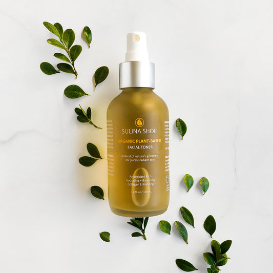Glass bottle of organic plant-based facial toner with white mist sprayer, surrounded by leaves on a clean, minimalist background.